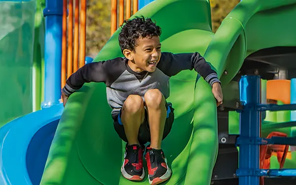 Boy riding a green curved playground slide, available from Bliss Products and Services.
