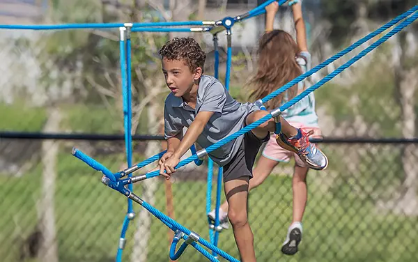 Children crossing a rope web traverse on synthetic turf, available from Bliss Products and Services.
