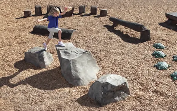 Child stepping across realistic climbing boulders in a nature play area with log and turtle steppers on engineered wood fiber, available from Bliss Products and Services.