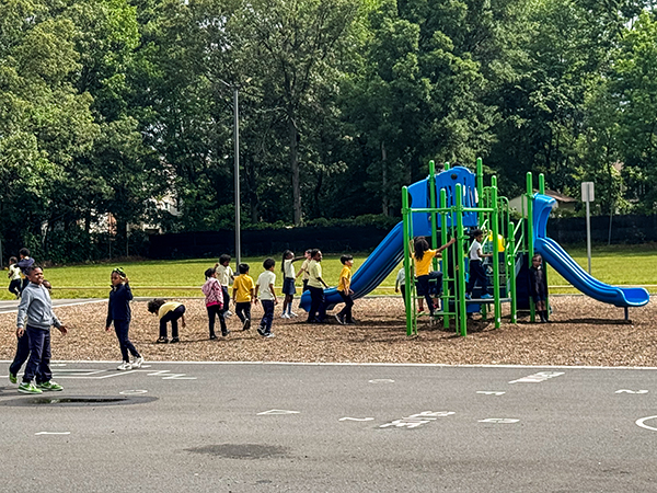 Wide view of the EXCEL Academy schoolyard with students playing on a blue and green playground structure and hard court games, installed by Bliss Products and Services in Maryland.