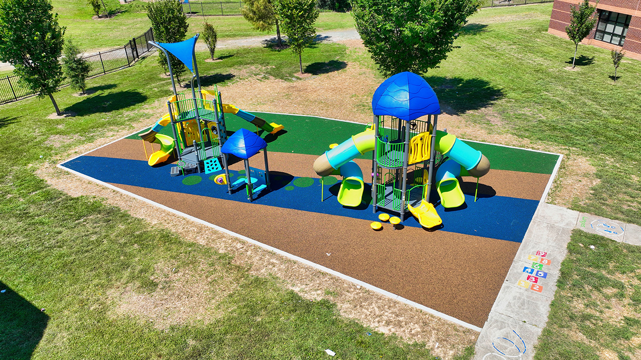 City of Charlotte Eastway playground aerial view with two play areas, poured in place safety surfacing in blue, green, and brown, a blue shade sail, and slide towers — Bliss Products and Services