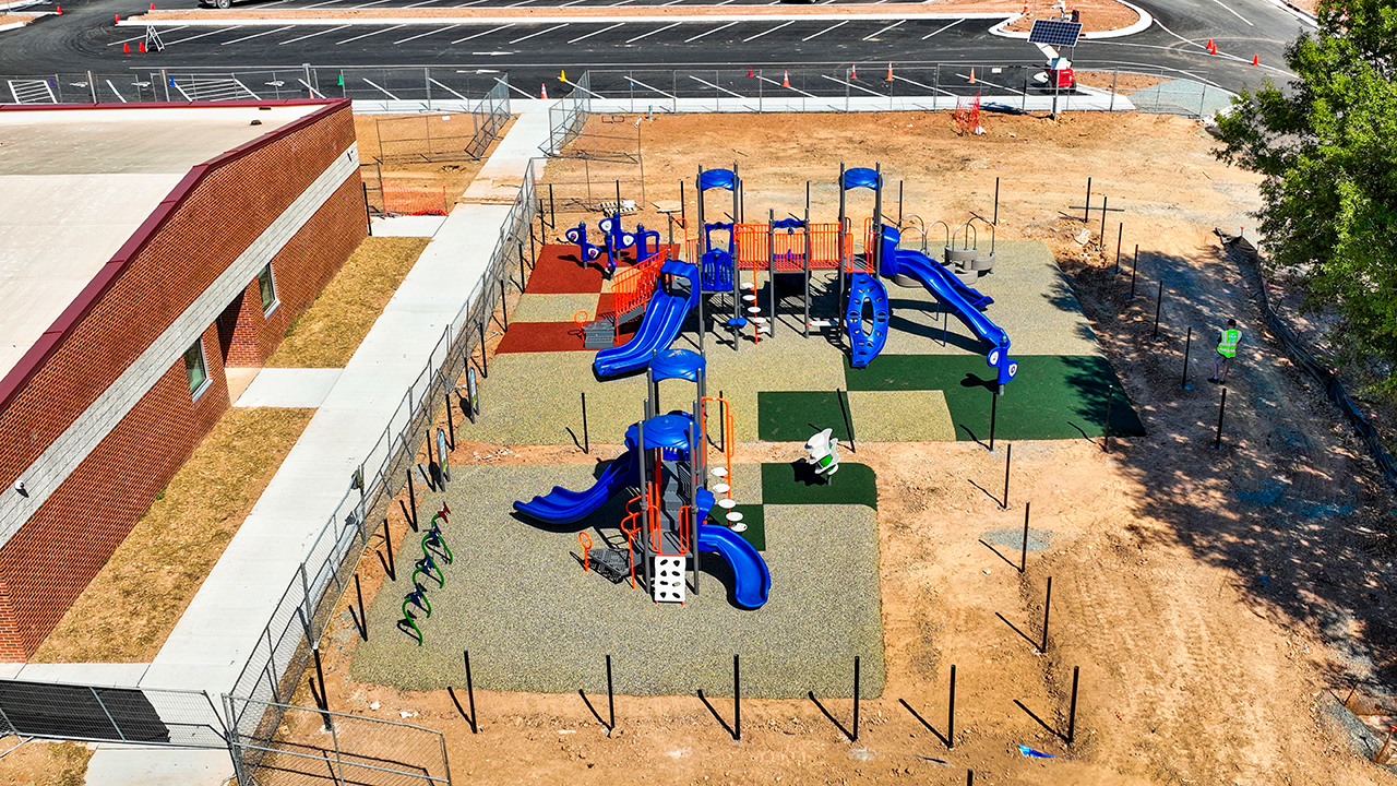 Glenn Elementary Playground | Bliss Products and Services Glenn Elementary playground aerial view with blue and orange play towers, slides, and poured in place safety surfacing in color blocks beside a brick school building — Bliss Products and Services