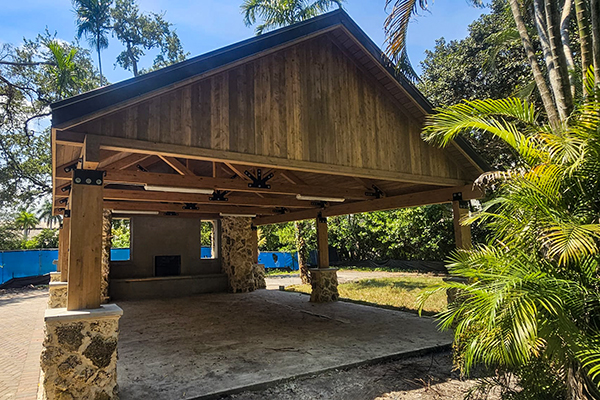 Side view of the Ben Butler Rock House Pavilion at Charles F. Vollman Park in Hollywood, Florida, showing timber framing, open-air shelter design, and preserved coral rock chimney. Bliss Products and Services.