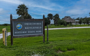 Entrance sign for Okeechobee Battlefield Historic State Park in Okeechobee County, Florida, with open grass, palm trees, and park buildings.