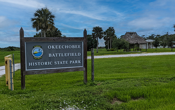 Entrance sign for Okeechobee Battlefield Historic State Park in Okeechobee County, Florida, with open grass, palm trees, and park buildings.