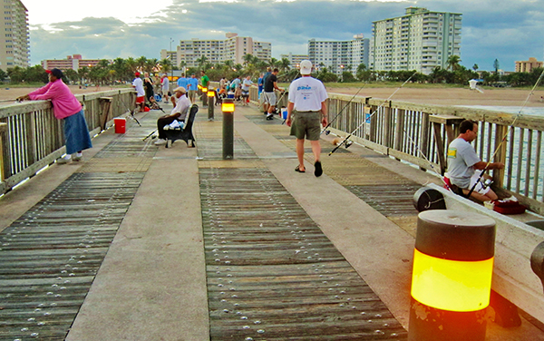 People fishing and walking on Pompano Beach Pier in Broward County, Florida, with beach and coastal buildings in the background.