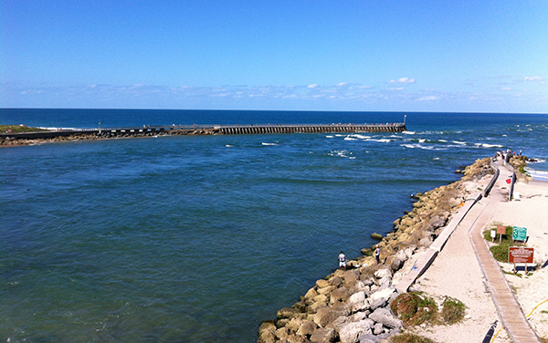 View of Sebastian Inlet in Florida, with the north jetty, blue water, shoreline, and Atlantic Ocean beyond.