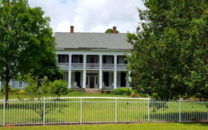 Historic two-story Chennault House with a long porch, mature trees, lawn, and white fence in Lincoln County, Georgia.